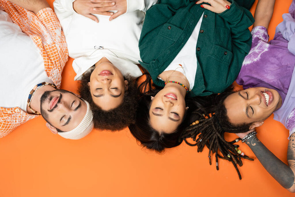 top view of smiling multiethnic friends in trendy clothes lying with closed eyes on orange background - Zdjęcie, obraz