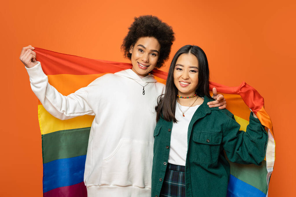 pozytywne i modne azjatyckie i afrykańskie amerykanki posiadające flagę Igbt izolowane na pomarańczowy - Zdjęcie, obraz