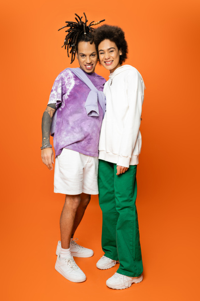 full length of cheerful african american woman and trendy man with dreadlocks smiling at camera on orange background - Foto, Imagem