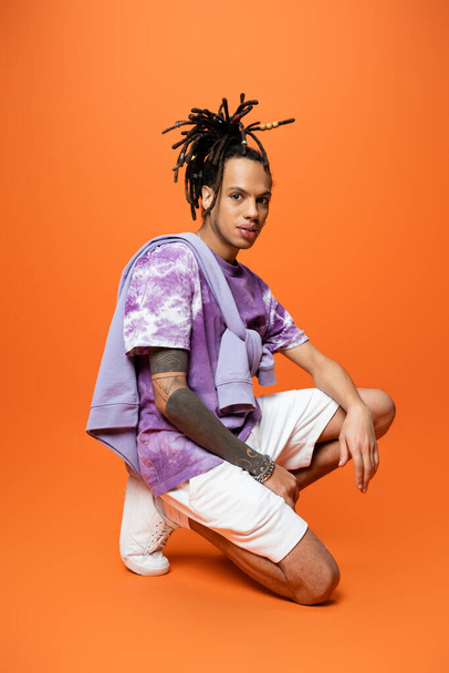 full length of multiracial man in trendy outfit sitting on haunches and looking at camera on orange background - Photo, Image