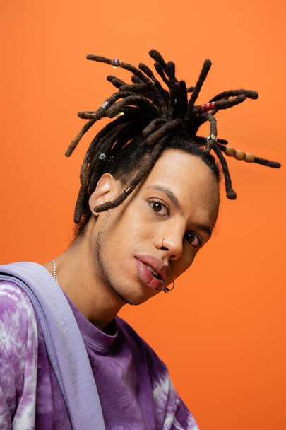 portrait of fashionable multiracial man with piercing and dreadlocks looking at camera isolated on orange - Photo, Image