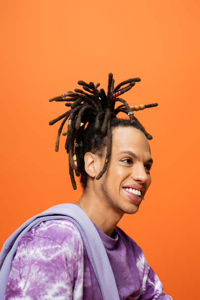 portrait of positive multiracial man with dreadlocks and piercing looking away isolated on orange - Photo, Image