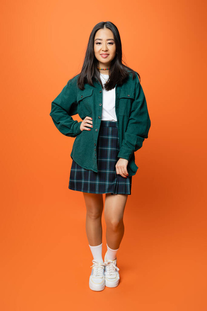full length of positive asian woman in green shirt and plaid skirt standing with hand on hip on orange background - Fotografie, Obrázek