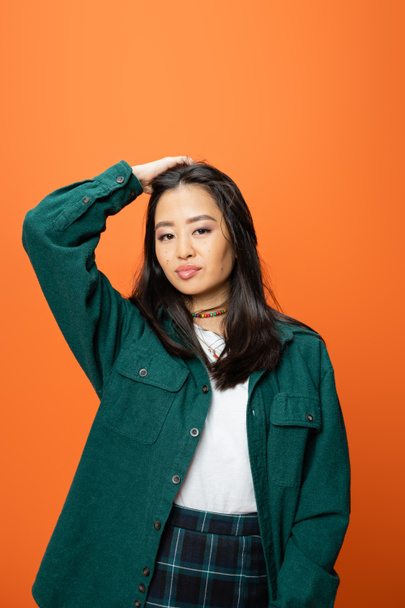 nuori aasialainen nainen vihreässä paidassa koskettaa ruskeaverikkö hiukset ja katsot kameraa eristetty oranssi - Valokuva, kuva