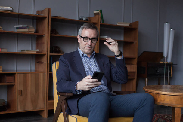 Elégant homme d'affaires âgé avec succès en costume bleu et lunettes regardant sur son téléphone portable contre le mur de bureau avec fond boockshelf - Photo, image