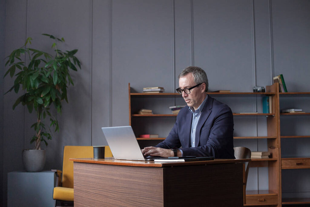 Starší zamyšlený podnikatel s šedými vlasy v brýlích a obleku pracuje na notebooku v kanceláři - Fotografie, Obrázek
