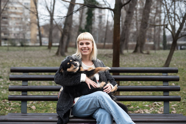 Young beautiful hipster woman sitting on a bench in the park with her adopted dog that saved his life from the street and the animal shelter and keeps him in her arms, gives him love food and care - Φωτογραφία, εικόνα
