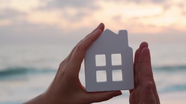 Батьки і дитина торкаються і демонструють білий будинок проти махаючого моря на заході сонця, створюючи форму будинку своїми руками
 - Кадри, відео