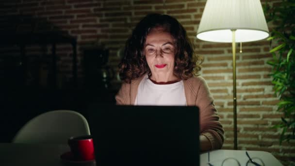 Mujer de mediana edad sonriendo confiado usando el ordenador portátil en casa - Metraje, vídeo
