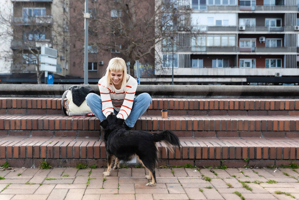 Νεαρή όμορφη γυναίκα hipster κάθεται στις σκάλες στο δρόμο με θετό σκυλί της υιοθέτησε από ένα καταφύγιο ζώων δίνοντάς του την αγάπη και την προσοχή - Φωτογραφία, εικόνα