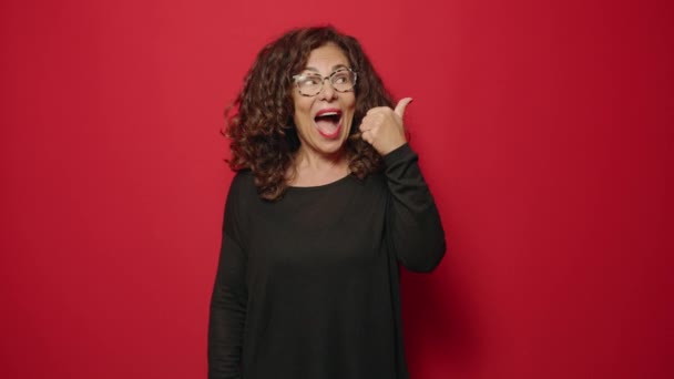 Mujer de mediana edad sonriendo confiada señalando con los dedos hacia un lado sobre el fondo rojo - Metraje, vídeo