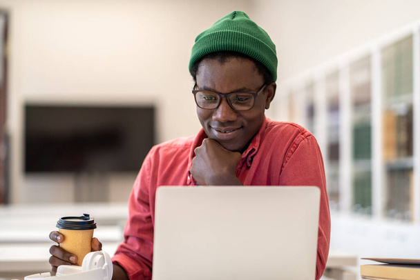 Pensieve Afro-Amerikaanse man met koffie in papieren beker gevoel tevreden met testresultaat, trots op projectwerk gedaan, houden kin ontspannen. Nadenken lachende zwarte man freelancer kijkt naar laptop scherm. - Foto, afbeelding