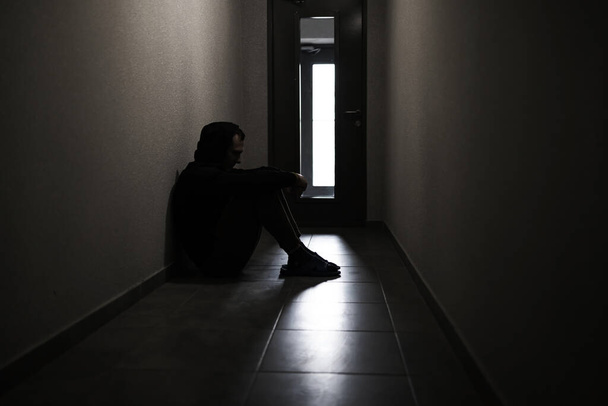 Η σιλουέτα ενός καταθλιπτικού άντρα που κάθεται στο διάδρομο μιας πολυκατοικίας. Θλιμμένος άνθρωπος, κλάμα, δράμα, μοναχική και δυστυχισμένη αντίληψη. - Φωτογραφία, εικόνα