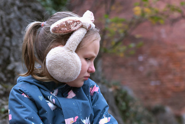 ένα κοριτσάκι με αφράτα ακουστικά στο φόντο της φθινοπωρινής φύσης με μια δυσαρεστημένη έκφραση στο πρόσωπό της, πορτραίτο προφίλ - Φωτογραφία, εικόνα