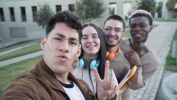 Group of positive multiracial friends in casual clothes smiling and showing peace gesture while taking selfie against building - Photo, Image