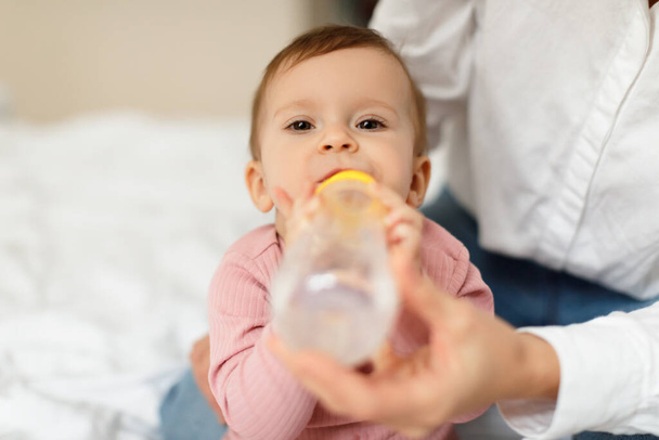 Mother giving her little daughter bottle of water feeding her sitting on bed at home, closeup shot, free space. Maternity leave, child care, parenting concept - Foto, afbeelding