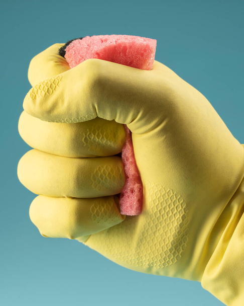 Guante de goma amarillo mano sostiene la esponja para lavar los platos y la limpieza, mano aprieta la esponja en el puño sobre fondo azul - Foto, imagen