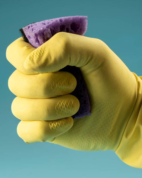 Hand in yellow rubber glove holds sponge for washing dishes and cleaning, hand clenches sponge into fist on blue background - Foto, Imagem