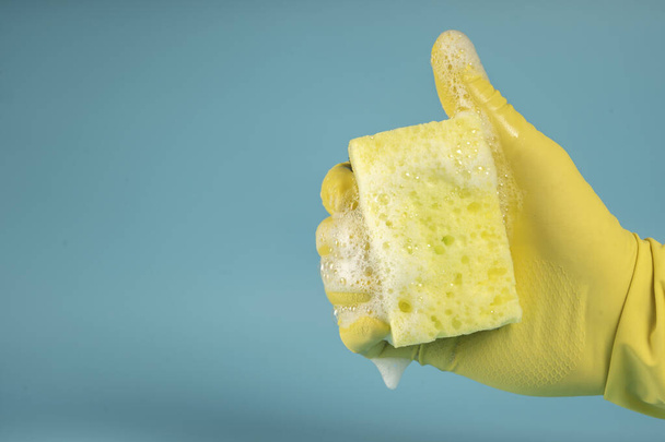 Hand in yellow rubber glove holds sponge for washing dishes and cleaning, hand clenches sponge into fist on blue background - Photo, Image