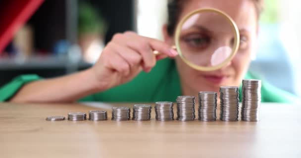 Businesswoman looks through magnifying glass at different stacks of coins on table. Symbol of financial analysis and search for maximum monetary profit - Footage, Video