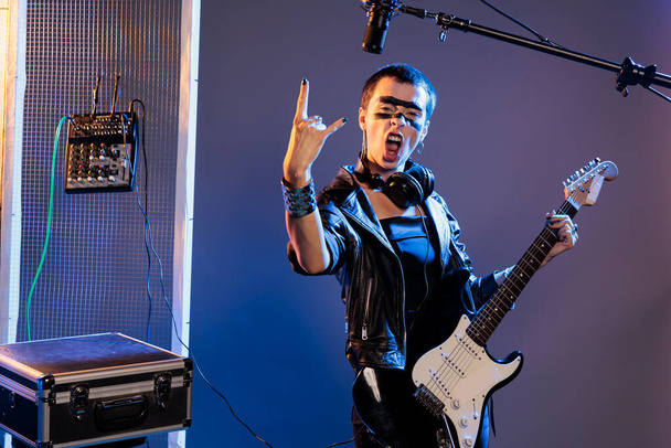 Musical artist doing rock sign and playing guitar, feeling ecstatic and doing solo concert in studio over background. Female singer showing rock and roll symbol at live show performance. - Fotó, kép