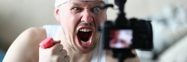 Portrait of screaming blogger man holding dumbbells at camera. Aggressive young sportsman filming video on digital camcorder for sports blog - Photo, Image