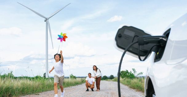 Concept of progressive happy family holding windmill toy and relax at wind farm with electric vehicle. Electric vehicle driven by clean renewable energy from wind turbine generator to charger station. - Photo, image