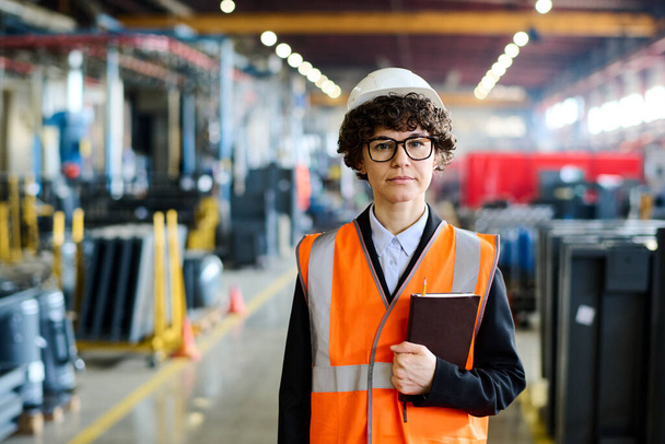 Young serious female factory manager in workwear and protective helmet looking at camera while standing in large workshop or warehouse - Foto, Bild
