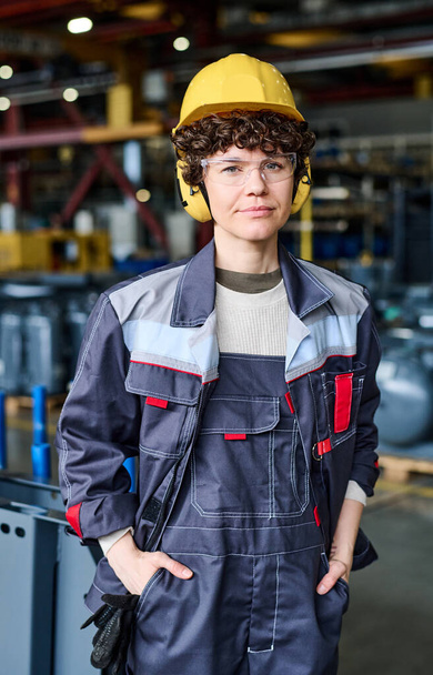 Young female worker of factory warehouse in uniform, protective helmet and headphones standing against new industrial equipment - Foto, afbeelding