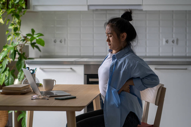 Unhealthy Asian woman suffering from backache while sits with laptop distant work or online education. Exhausted Korean freelancer girl have acute lower back pain from incorrect posture on bad chair. - Photo, Image