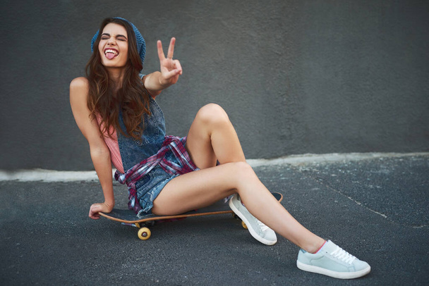 I wont fall today I promise. Portrait of a cheerful young woman seated on a skateboard while showing the peace sign outside during the day - Foto, imagen