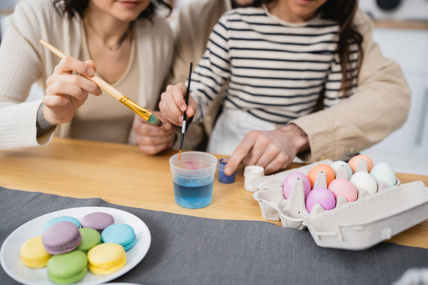 Cropped view of family holding paintbrushes near paints and Easter eggs in kitchen  - 写真・画像