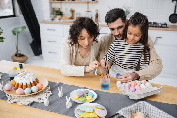 Familia para colorear huevos de Pascua cerca de pastel y galletas en la cocina  - Foto, Imagen