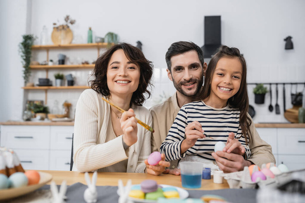 Smiling family looking at camera while coloring Easer eggs in kitchen  - Photo, image