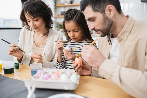 концентрированная девушка рисует пасхальное яйцо рядом с родителями на кухне  - Фото, изображение