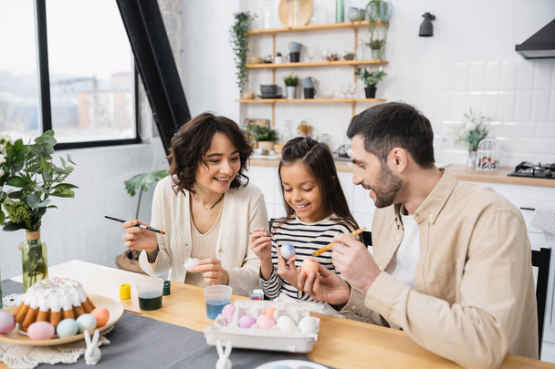 Smiling parents and child coloring eggs near Easter cake at home  - Photo, Image