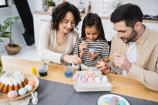 Smiling woman coloring Easter egg near family and food at home - Photo, image