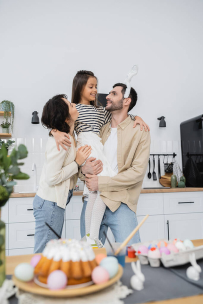 Parents holding daughter near blurred Easter cake and eggs at home  - Φωτογραφία, εικόνα