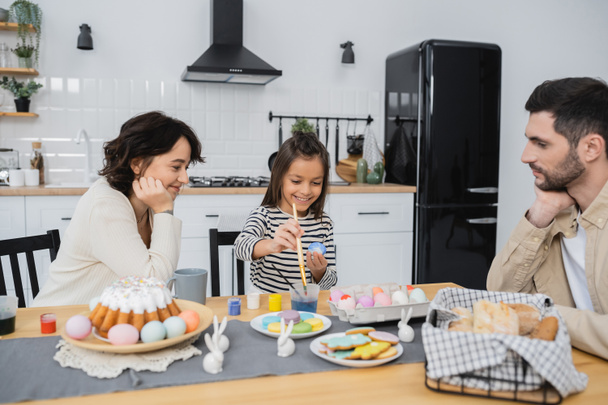 Smiling girl coloring Easter eggs near parents and food in kitchen  - Foto, Bild