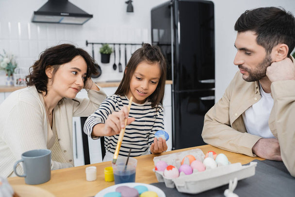 Parents looking at happy daughter coloring Easter eggs near macaroons at home  - Fotó, kép