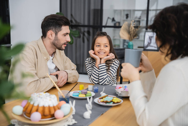 Веселый ребенок сидит рядом с родителями и вкусная пасхальная еда дома  - Фото, изображение