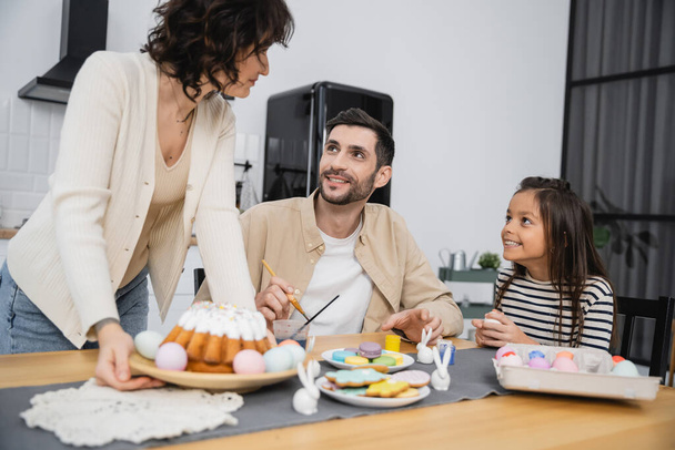 Улыбающаяся семья раскрашивает яйца рядом с женщиной мурлыкая тарелка с пасхальным тортом на столе дома  - Фото, изображение