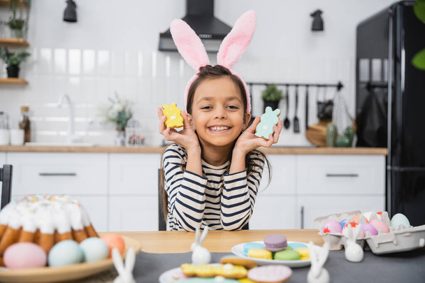 Cheerful kid in headband holding cookies near Easter cake and eggs at home  - Photo, image