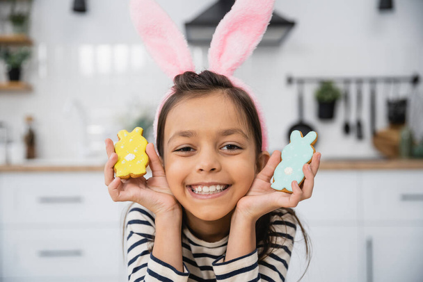 Улыбающийся ребенок в пасхальном повязке с кроличьими ушами, держащий печенье на кухне  - Фото, изображение