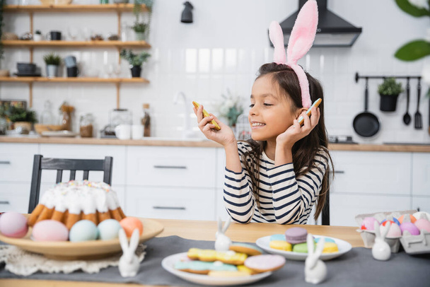 Улыбающийся ребенок в повязке смотрит на пасхальное печенье рядом с едой на кухне  - Фото, изображение