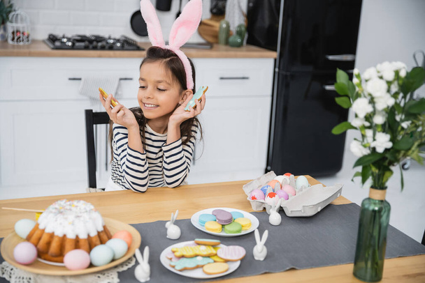Happy preteen kid in headband holding Easter cookies near flowers and food at home  - Fotografie, Obrázek