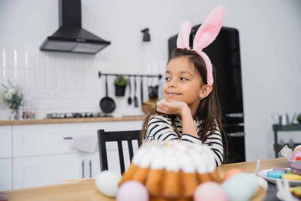 Улыбающийся ребенок в повязке с кроличьими ушами возле пасхального торта дома  - Фото, изображение