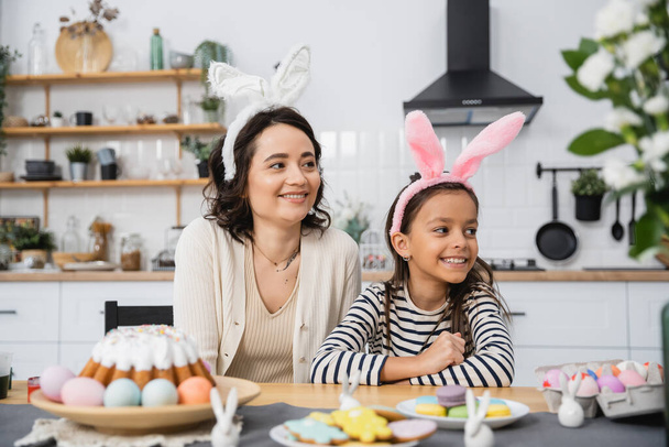 Mosolygó anya és gyerek nyuszifül fejpánt ül közel Húsvéti torta és tojás otthon  - Fotó, kép