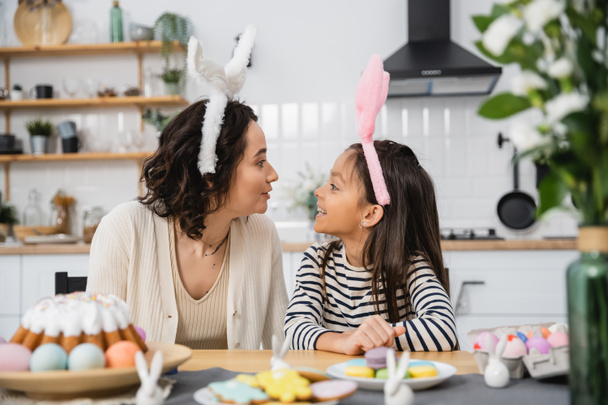 Side view of smiling mom and kid in headbands sitting near Easter food at home  - Фото, зображення