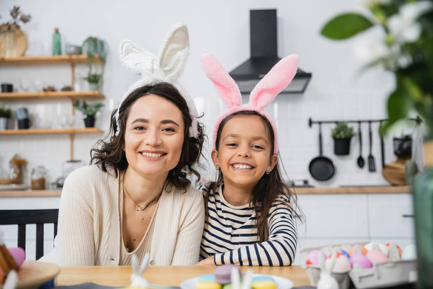 Smiling mom and kid in Easter headbands looking at camera near eggs and macaroons at home  - Foto, imagen